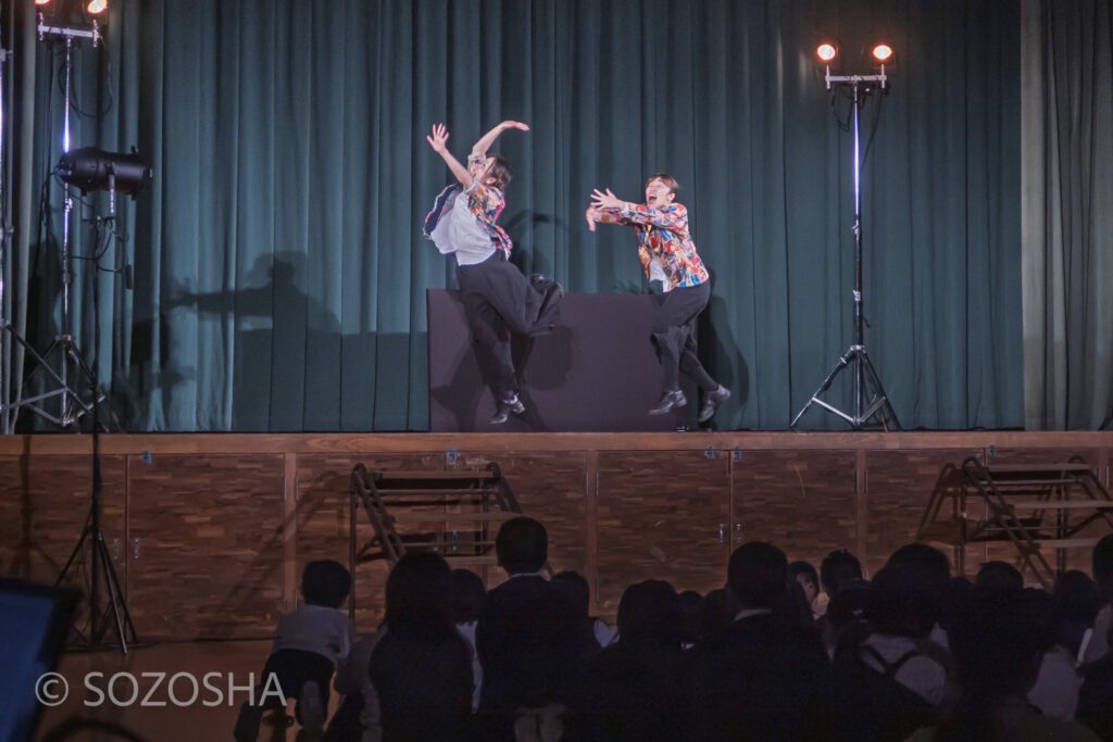 ステージ上で二人の俳優が大きく跳ね上がるように動き、体育館の児童たちが見上げている。 | 芸術鑑賞会 | パントマイム |マイムボックス〜おしゃべりなカラダ〜 | いいむろなおき